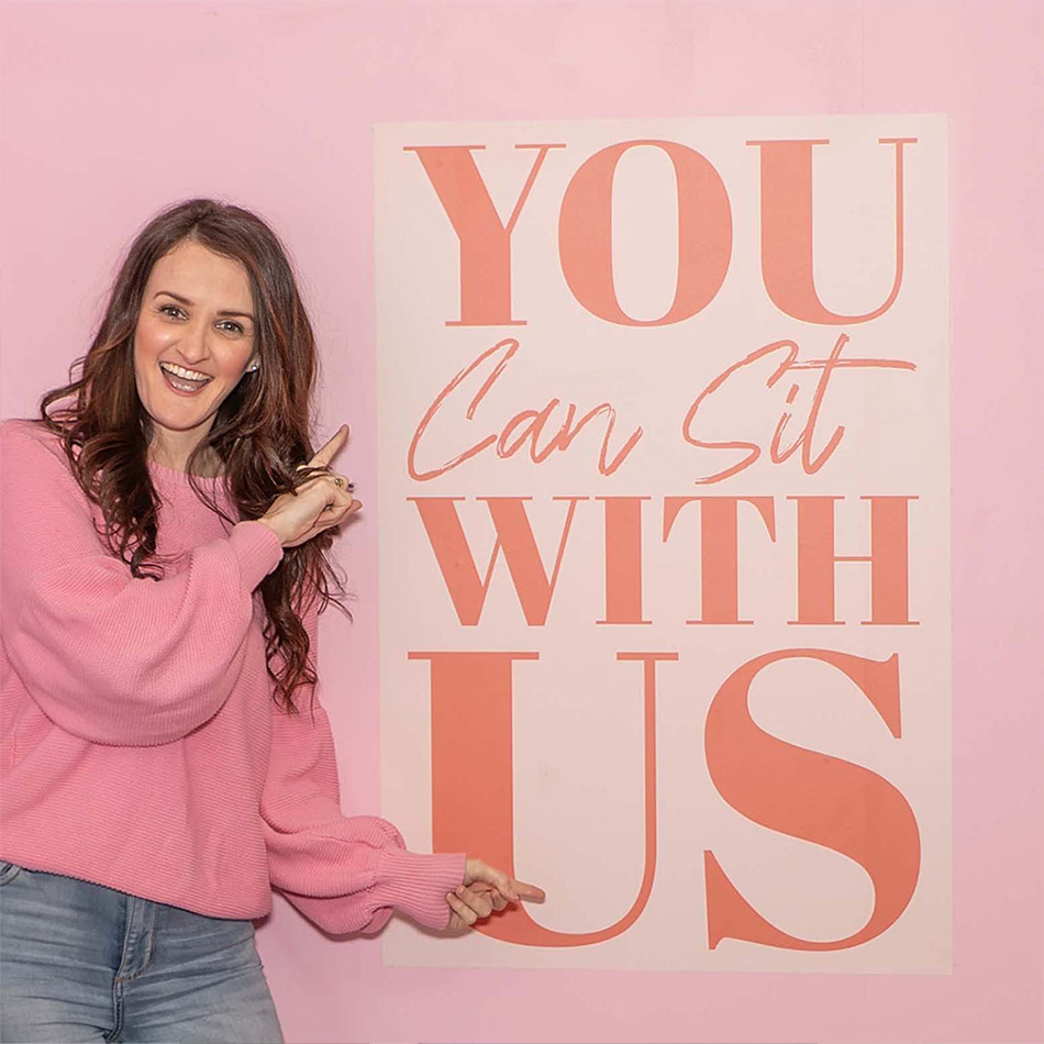 Brunette woman standing in front of Kindness sign that says 'You Can Sit With Us' and pink wall.